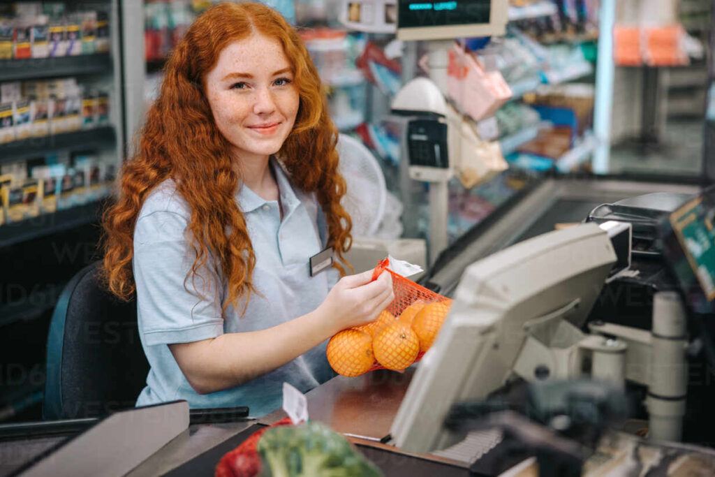 Woman cashier sitting behind checkout and working in supermarket. Female checkout counter clerk scanning grocery products and looking at camera.